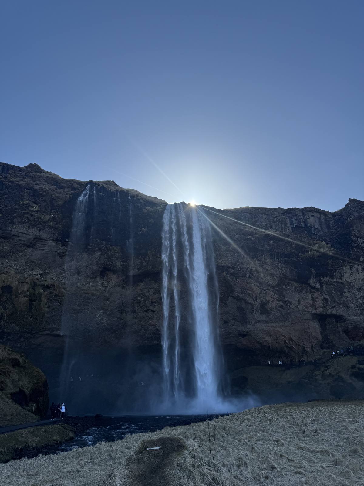 Seljalandsfoss waterfall dropping off a green cliff on Iceland's South Coast, the sun flaring behind the falls